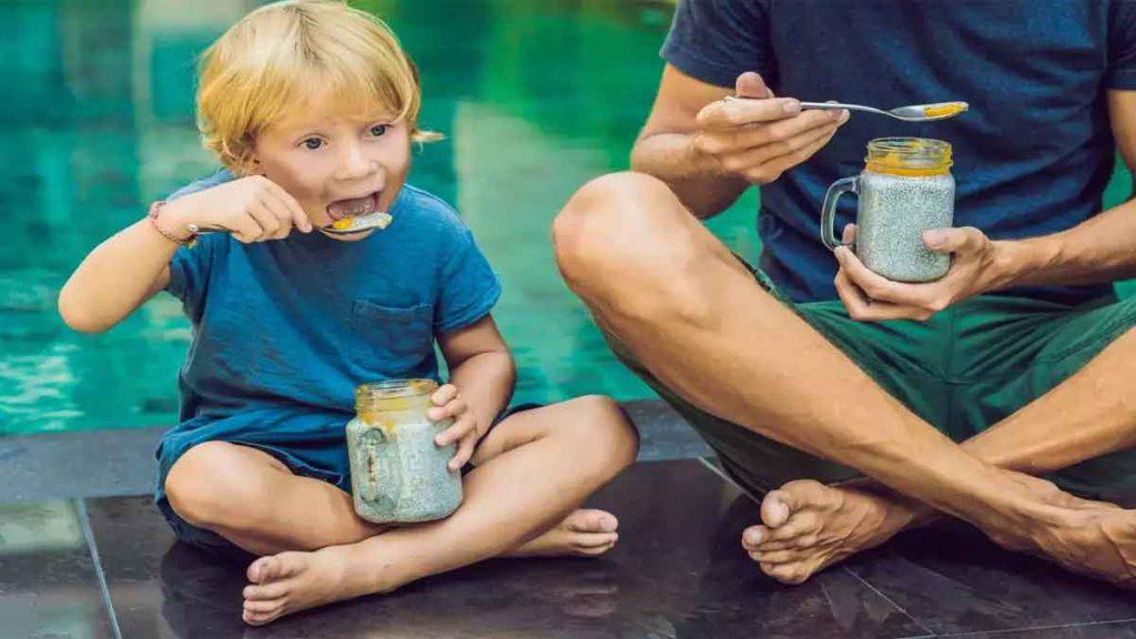 یک پدر و کودک که در بیرون نشسته اند و هر کدام یک مخلوط یک لیوان مخلوط دانه چیا با میوه در دست دارند و مشغول خوردن آن هستند و به خواص دانه چیا در کودکان اشاره دارد