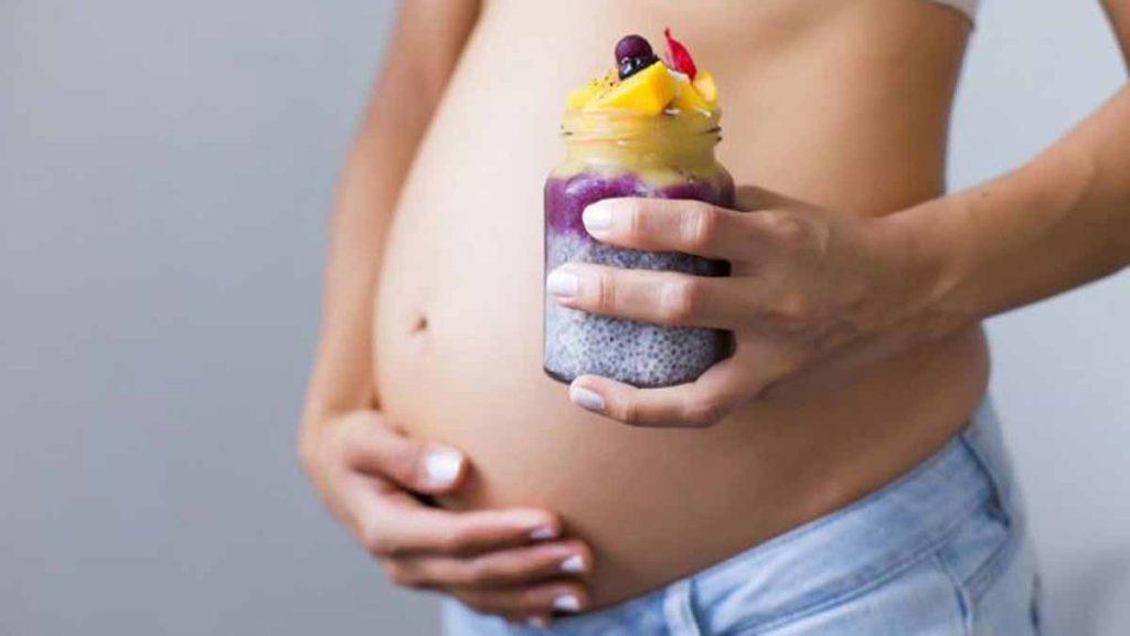 یک خانم باردار که دست راستش را در زیر شکمش قرار داده و در دست چپش یک لیوان که مخلوط دانه چیا با میوه های مختلف است قرار دارد و به خواص دانه چیا در بارداری اشاره دارد