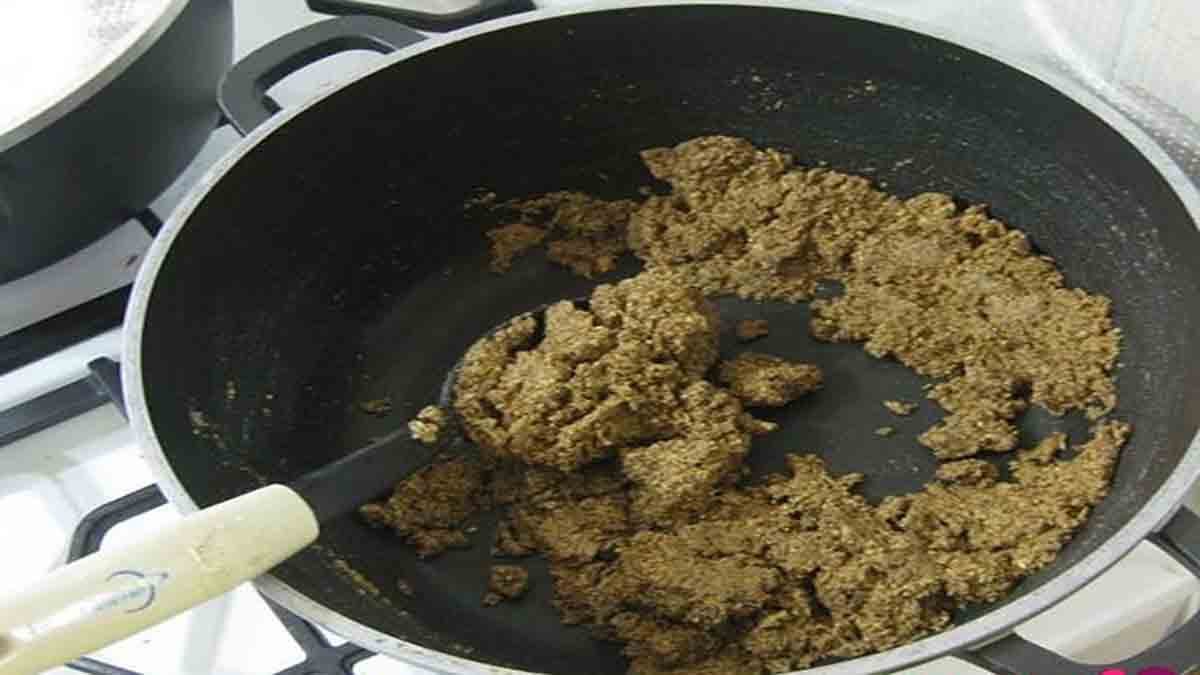 مرحله دوم پخت فسنجون با مرغ و تفت گردو و پیاز یک ماهی تابه حاوی گردو آسیاب شده و پیاز که بر روی گاز قرار دارد و قاشقی در آن قرار دارد و مرحله دوم بخت فسنجون با مرغ و تفت گردو و پیاز را نشان می دهد